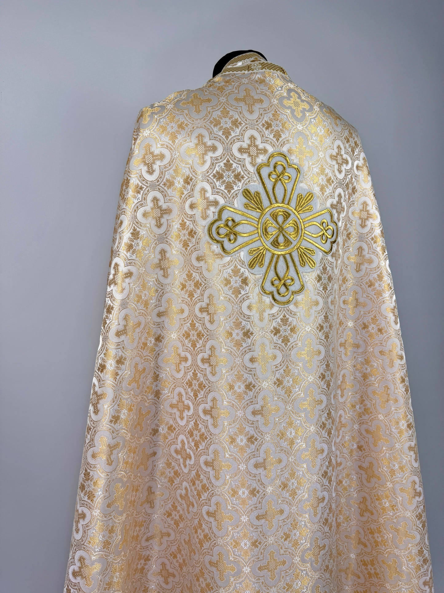Ecclesiastical vestment with intricate lace design and gold cross emblem on a gray background