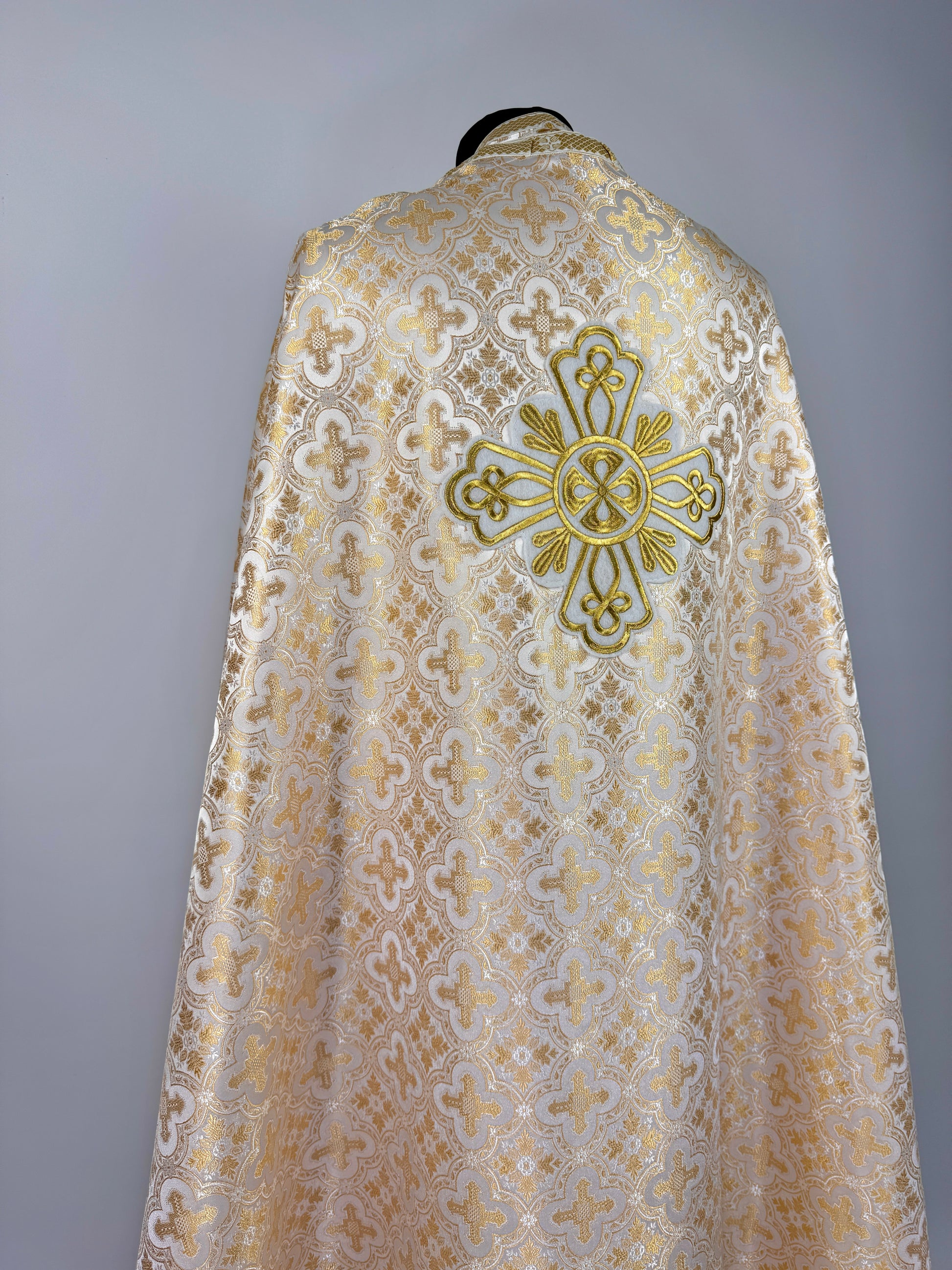 Ecclesiastical vestment with intricate lace design and gold cross emblem on a gray background